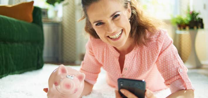 Portrait of smiling modern woman in blouse and white pants with piggy bank searching for carpet cleaning service on a smartphone while laying on white carpet at modern home in sunny day.