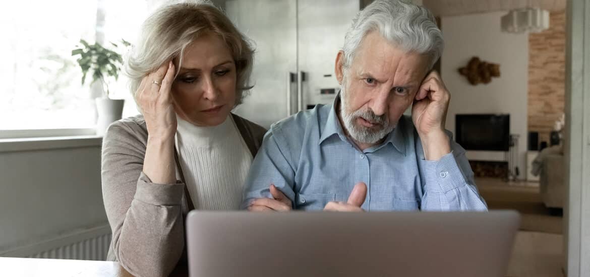 Upset 60s senior couple getting shocking bad news, using laptop together at home, reading email message or talking via video call. Frustrated elder retired husband and wife looking at computer screen