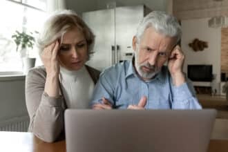 Upset 60s senior couple getting shocking bad news, using laptop together at home, reading email message or talking via video call. Frustrated elder retired husband and wife looking at computer screen