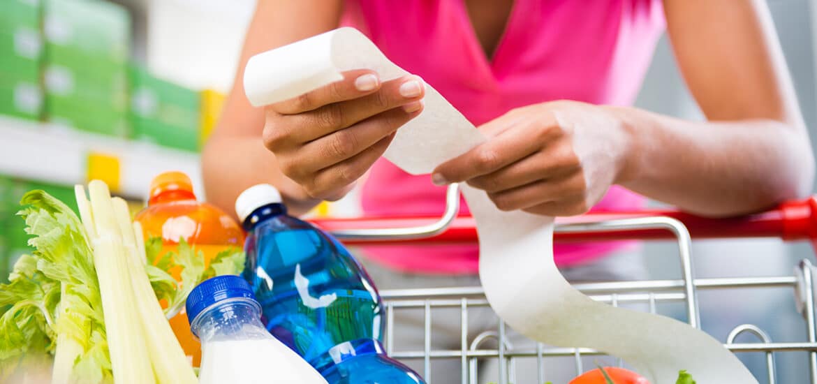 Unrecognizable woman checking a long grocery receipt leaning to a full shopping cart at store.