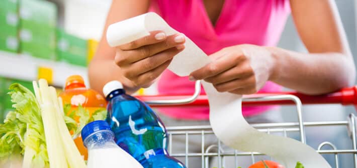 Unrecognizable woman checking a long grocery receipt leaning to a full shopping cart at store.