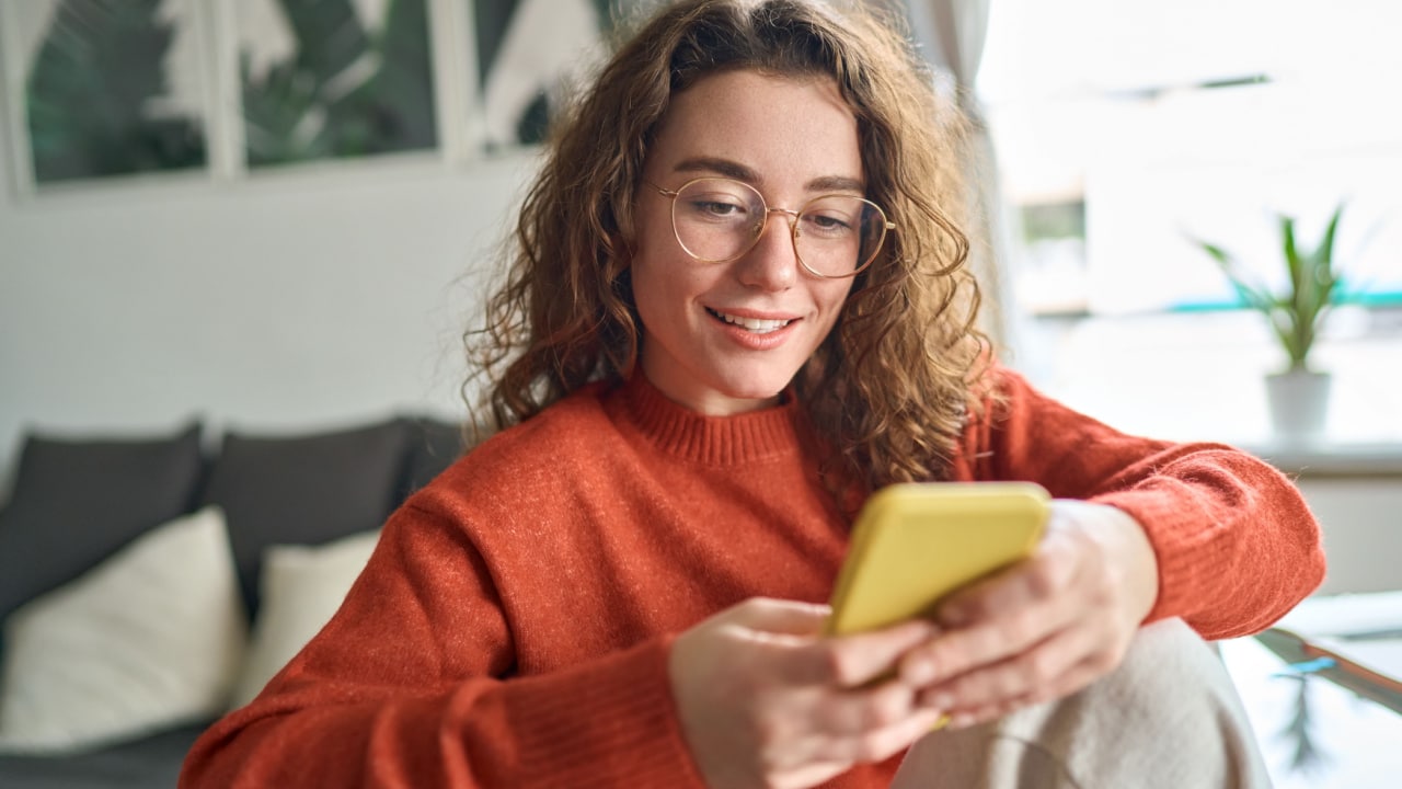 Young smiling woman wearing glasses holding smartphone using cellphone to study gardening tips online